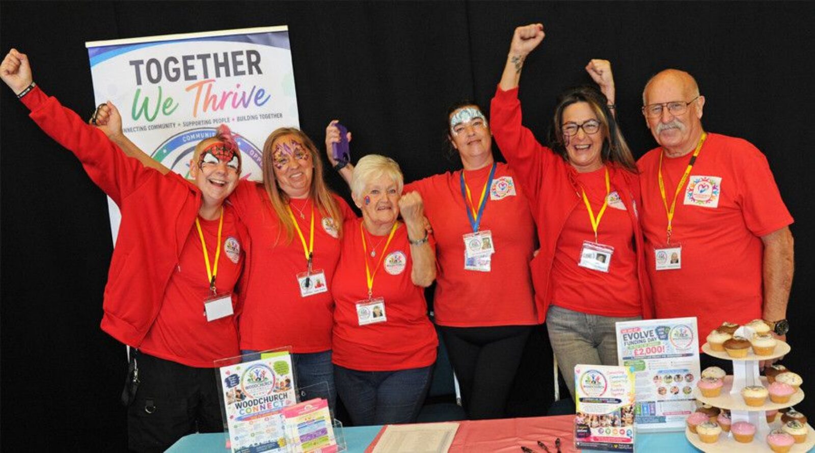A mixed group of six people with red tops, arms aloft, in front of a table with cakes and leaflets about an Evolve Woodchirch Connects event and a banner in the background headed Together We thrive.